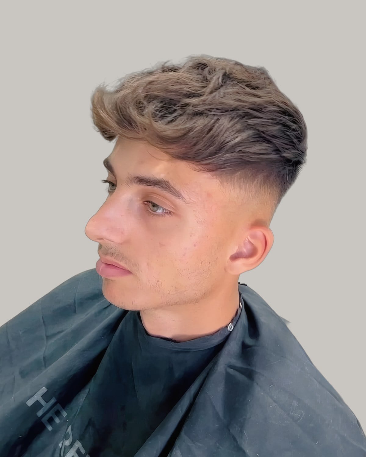Man in a barber's cape with a light brown textured fringe over a high skin fade, seen in three-quarter profile