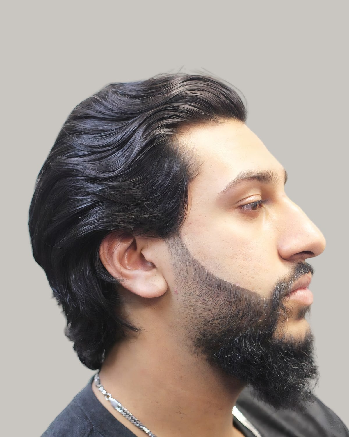 Bearded man with black, wavy hair slicked back to the nape and a silver chain, seen in profile