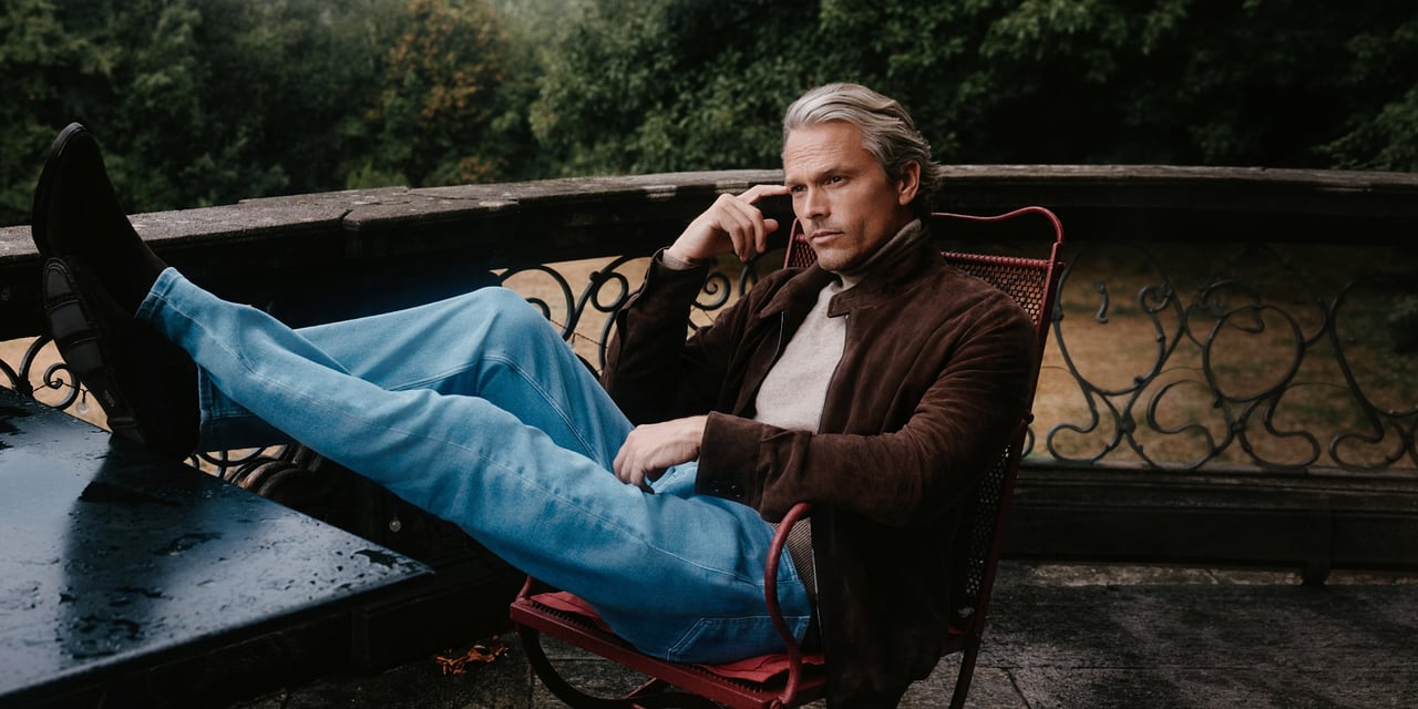Man with swept-back silver hair, in a brown suede jacket and jeans, sitting with his feet up on a terrace table