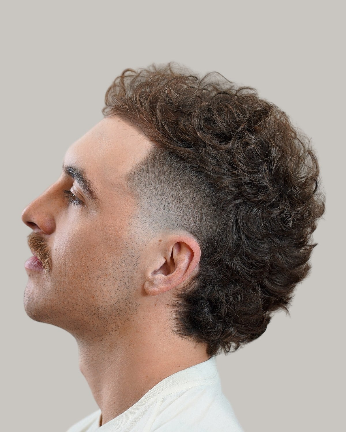 Man with a mustache and a brown curly mullet with a skin fade, seen in profile