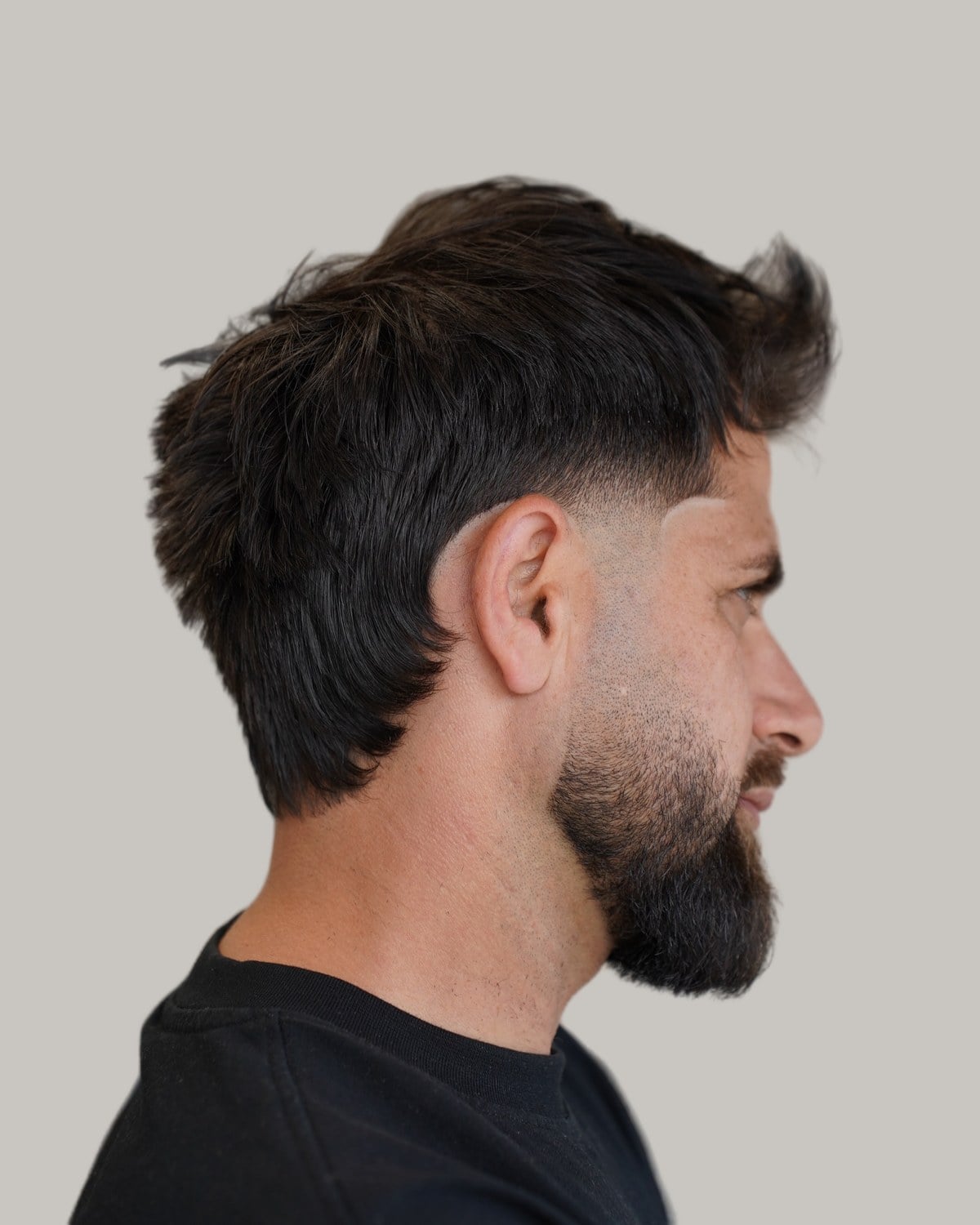 Bearded man with a dark textured mullet and a low fade, seen in profile