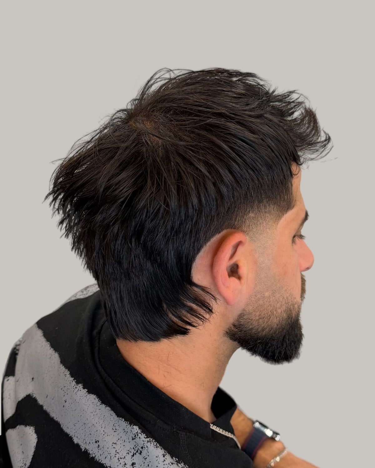 Bearded man with a dark textured mullet and a low taper, seen from behind at an angle