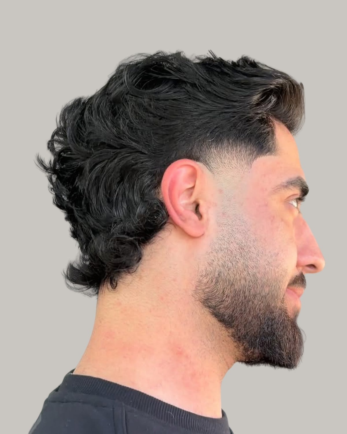 Bearded man with a dark wavy mullet and a low taper, seen in profile