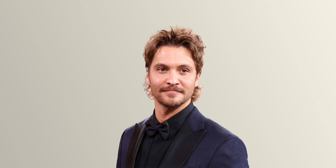 Man with a tousled light brown modern mullet and a short beard, in a navy tuxedo and black bow tie