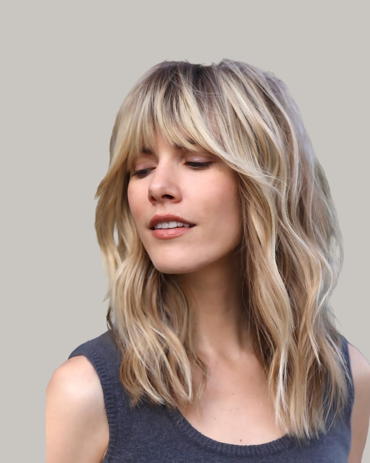 Woman in a gray tank top with blonde waves and curtain bangs, eyes lowered