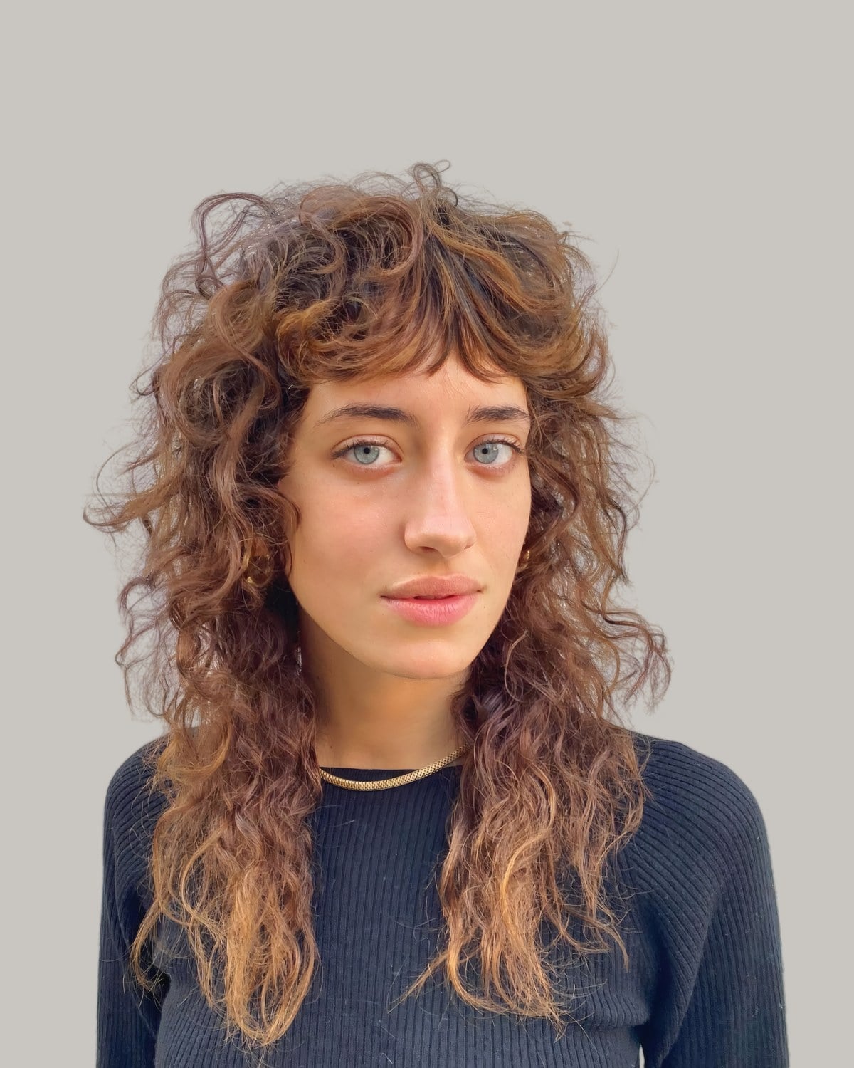 Woman in a black sweater and a gold necklace with a long, light brown curly shag and curly bangs