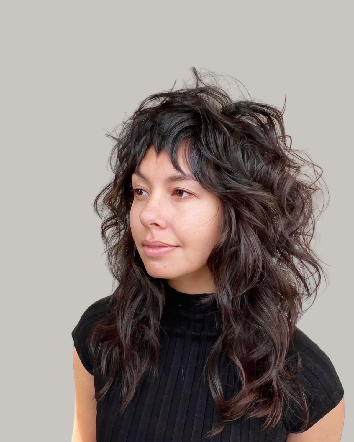 Woman in a black ribbed top with a long, dark shag of loose curls and wispy bangs, looking to the side