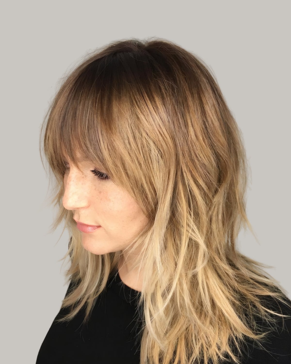 Woman with freckles and a long blonde shag with darker roots and wispy bangs, looking down