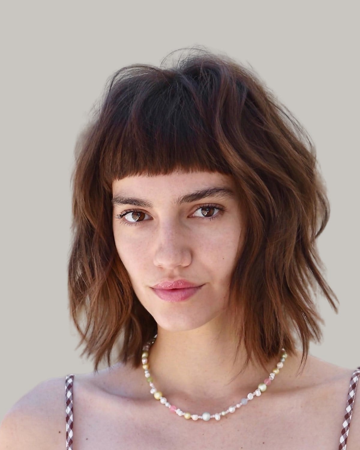 Woman in a gingham top and a beaded necklace with a brown shaggy bob and short, blunt bangs