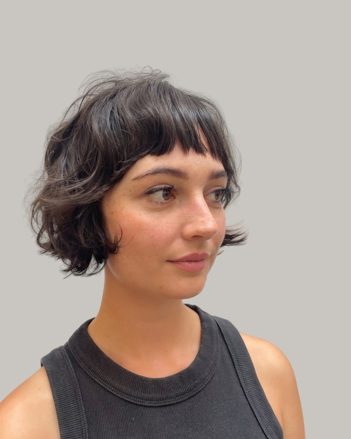 Woman in a grey tank top with a dark brown, chin-length wavy shag and micro bangs, looking to the side