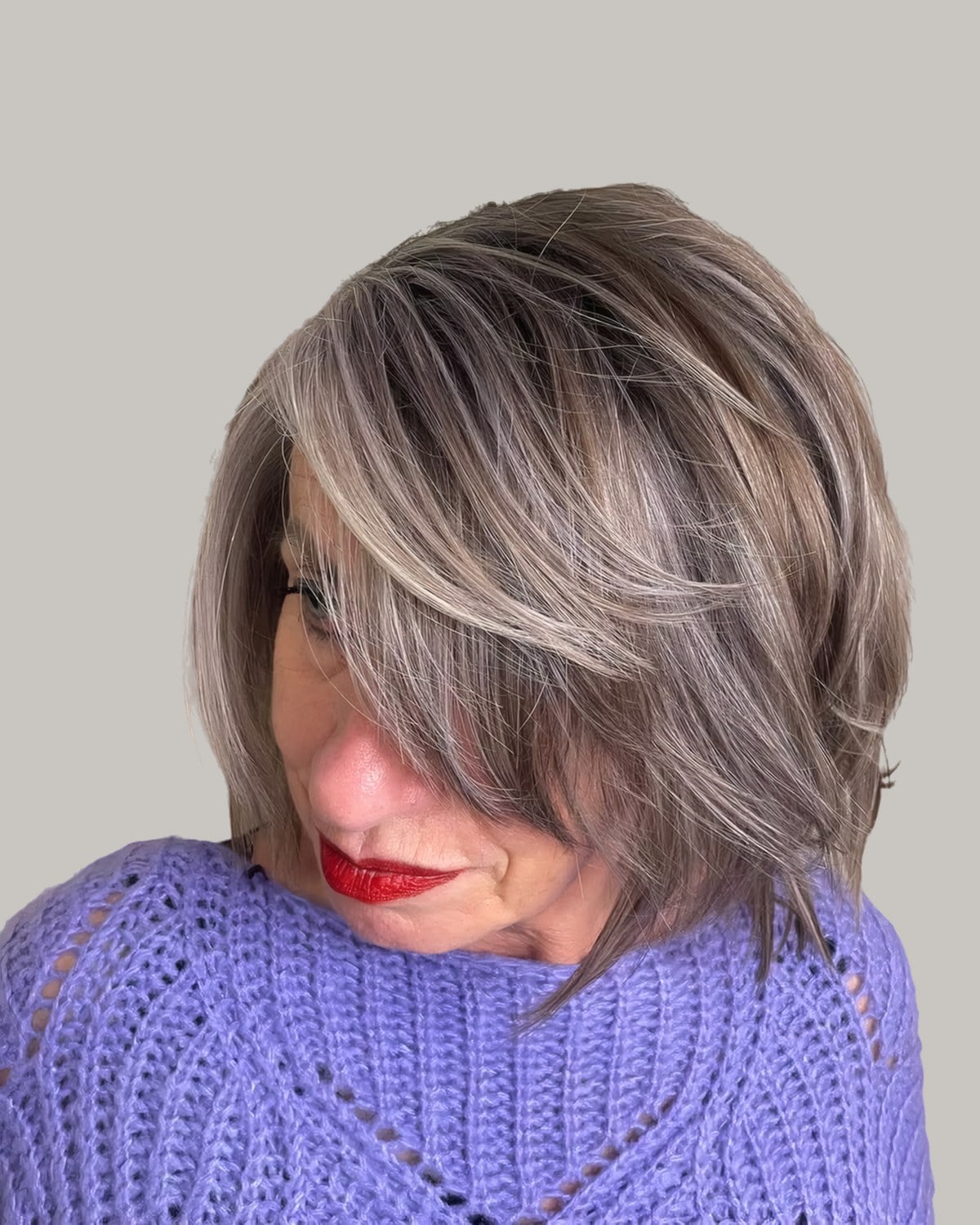 Woman in a purple sweater and red lipstick looking down, brown shaggy bob with silver highlights