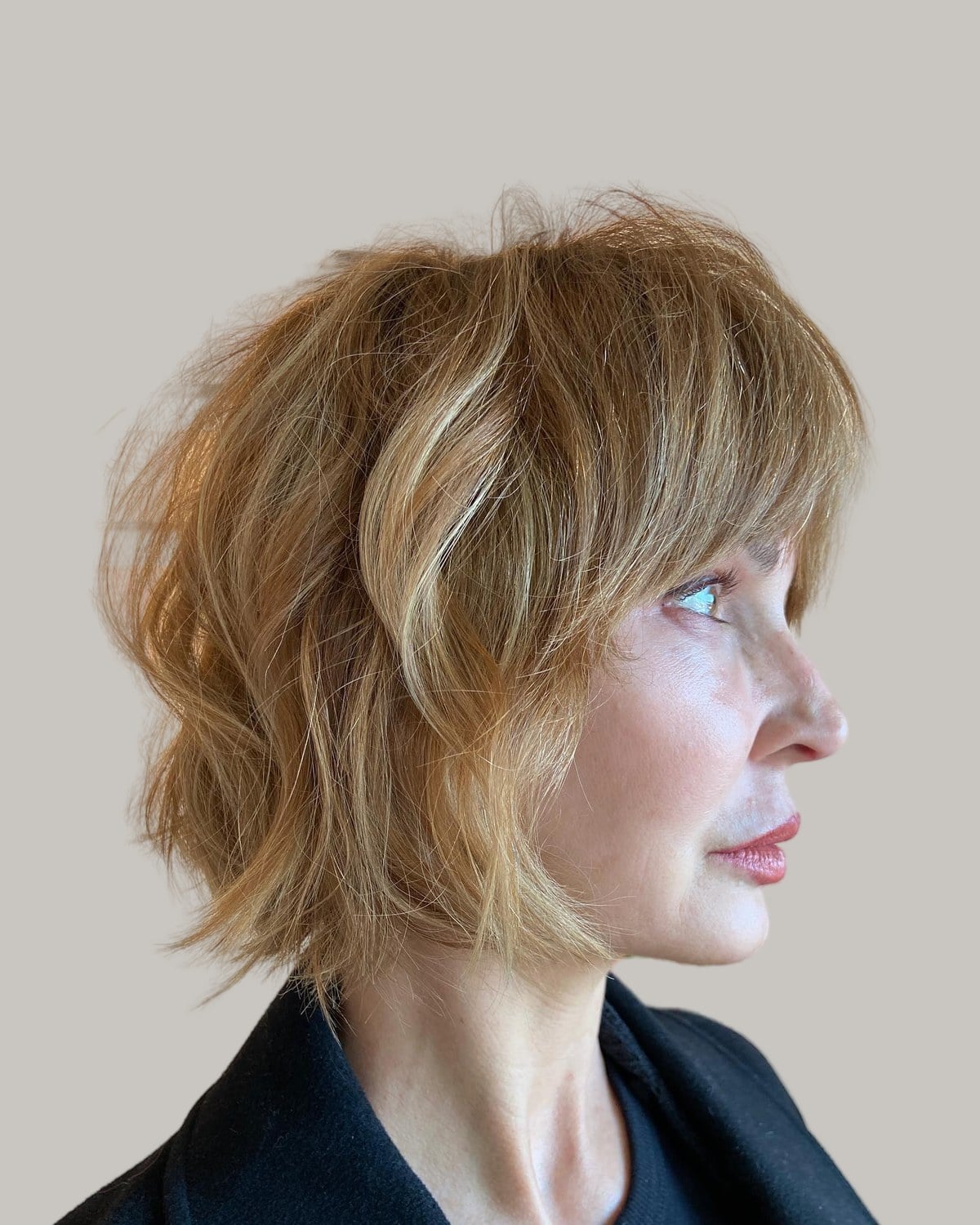 Woman in a black coat seen in profile, honey-blonde short shaggy bob with soft bangs