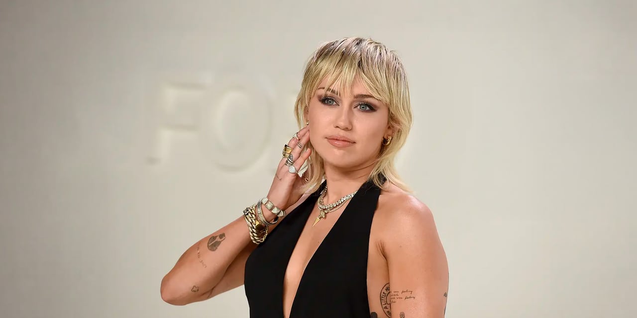 Woman with a blonde shag and wispy bangs, in a black halter dress, one hand raised to her hair
