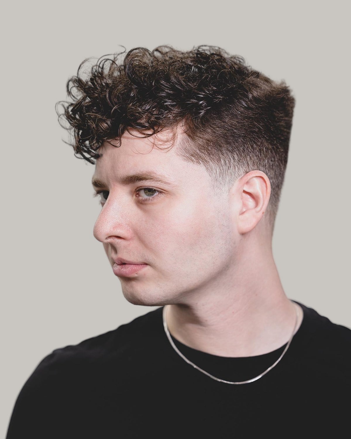 Young man with a curly top and a mid taper fade, wearing a silver chain, seen at an angle