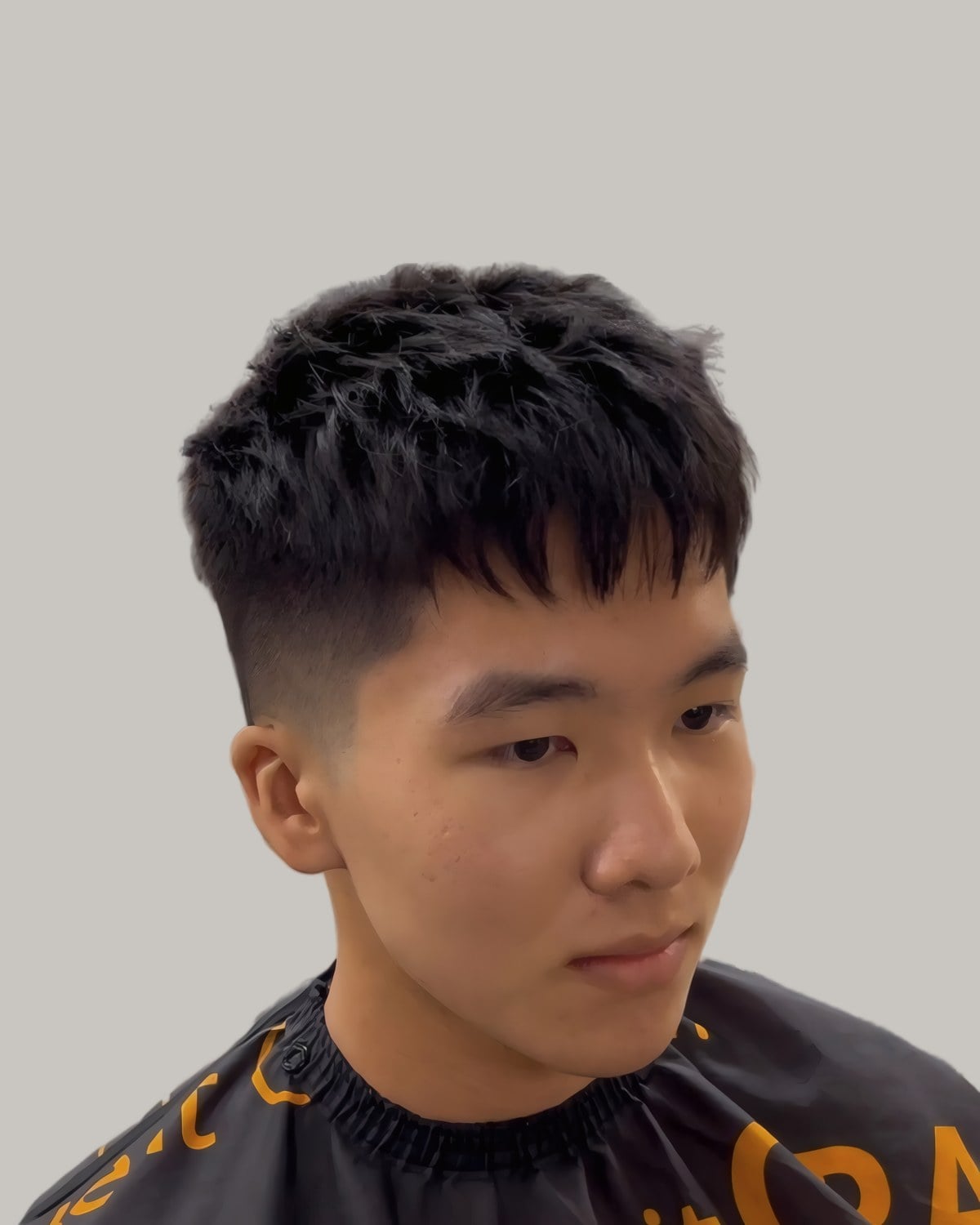 Young Asian man with a choppy textured crop and a low fade, in a barber's cape