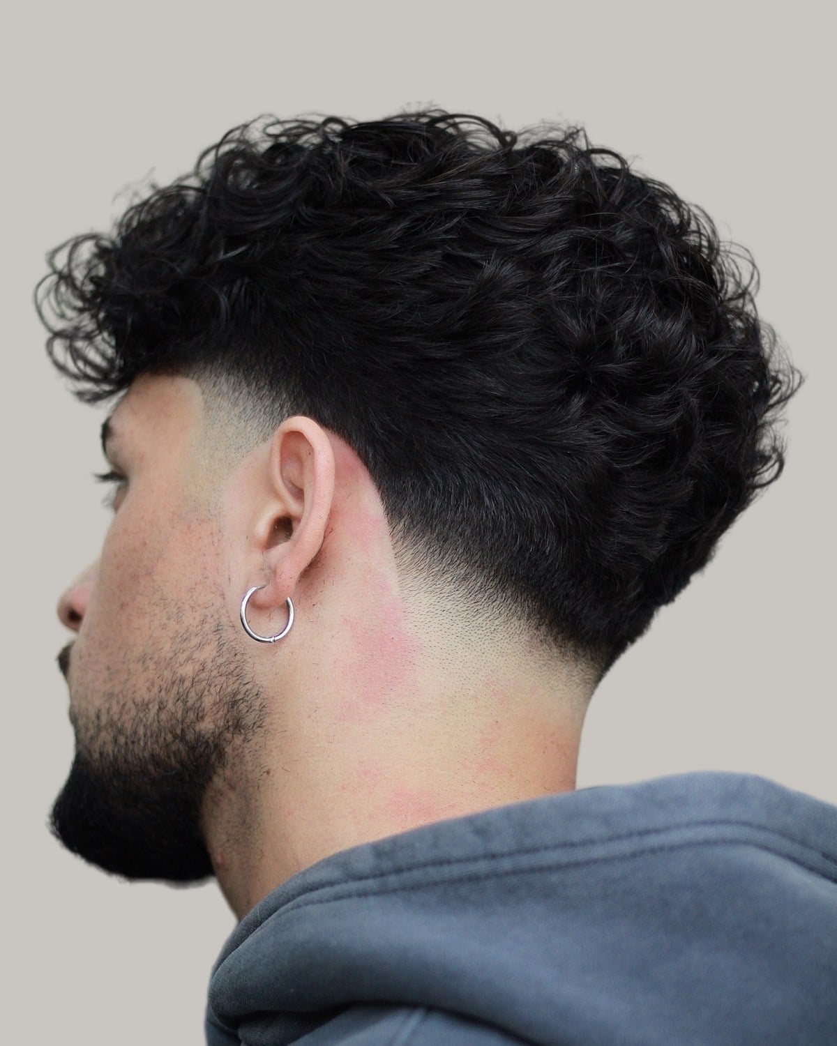 Bearded man with a dense black curly top, a blowout taper and a hoop earring, seen from the side and behind
