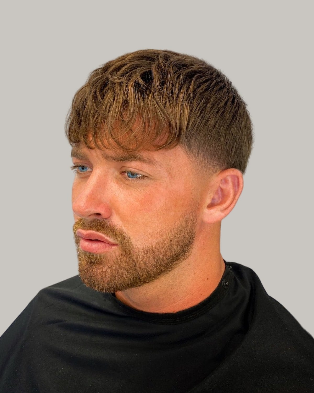 Bearded man with a heavy auburn-brown fringe and a soft taper at the sides, looking to the side