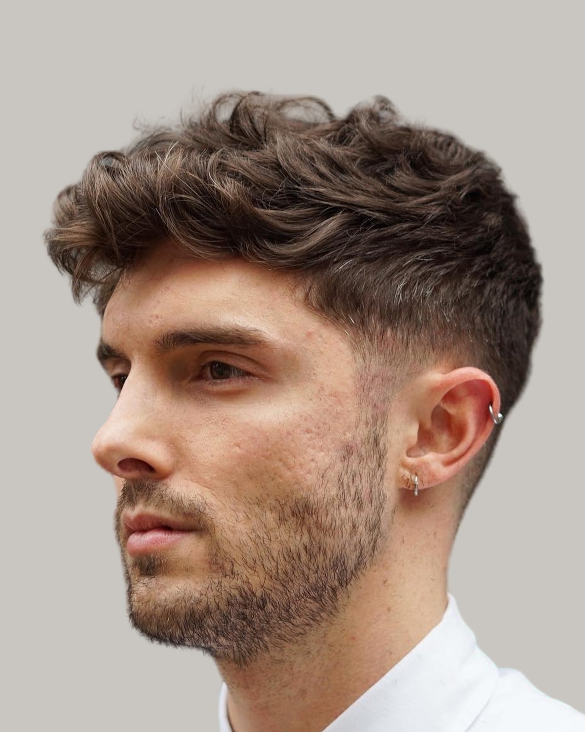 Man with a short beard and wavy brown hair pushed forward into a fringe, with a low taper, seen at an angle