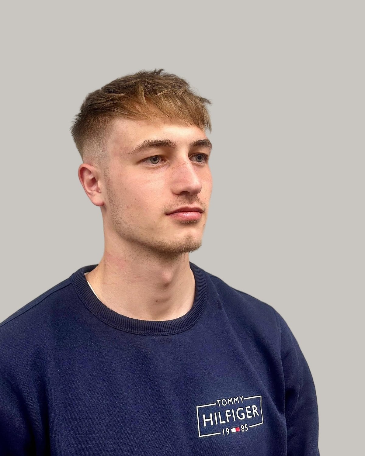 Young man with a light brown messy fringe and a mid taper fade, wearing a navy sweatshirt, seen at an angle