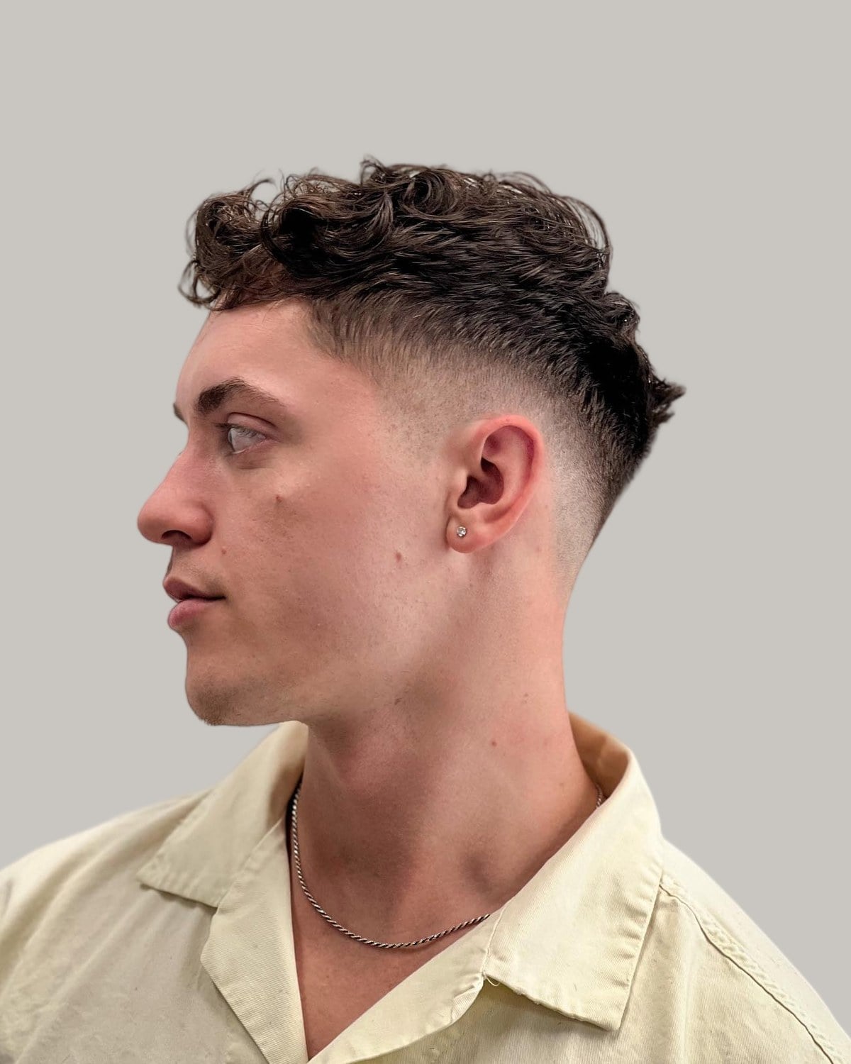 Man in a cream shirt and chain with a wavy textured top and a mid blowout taper, seen in profile