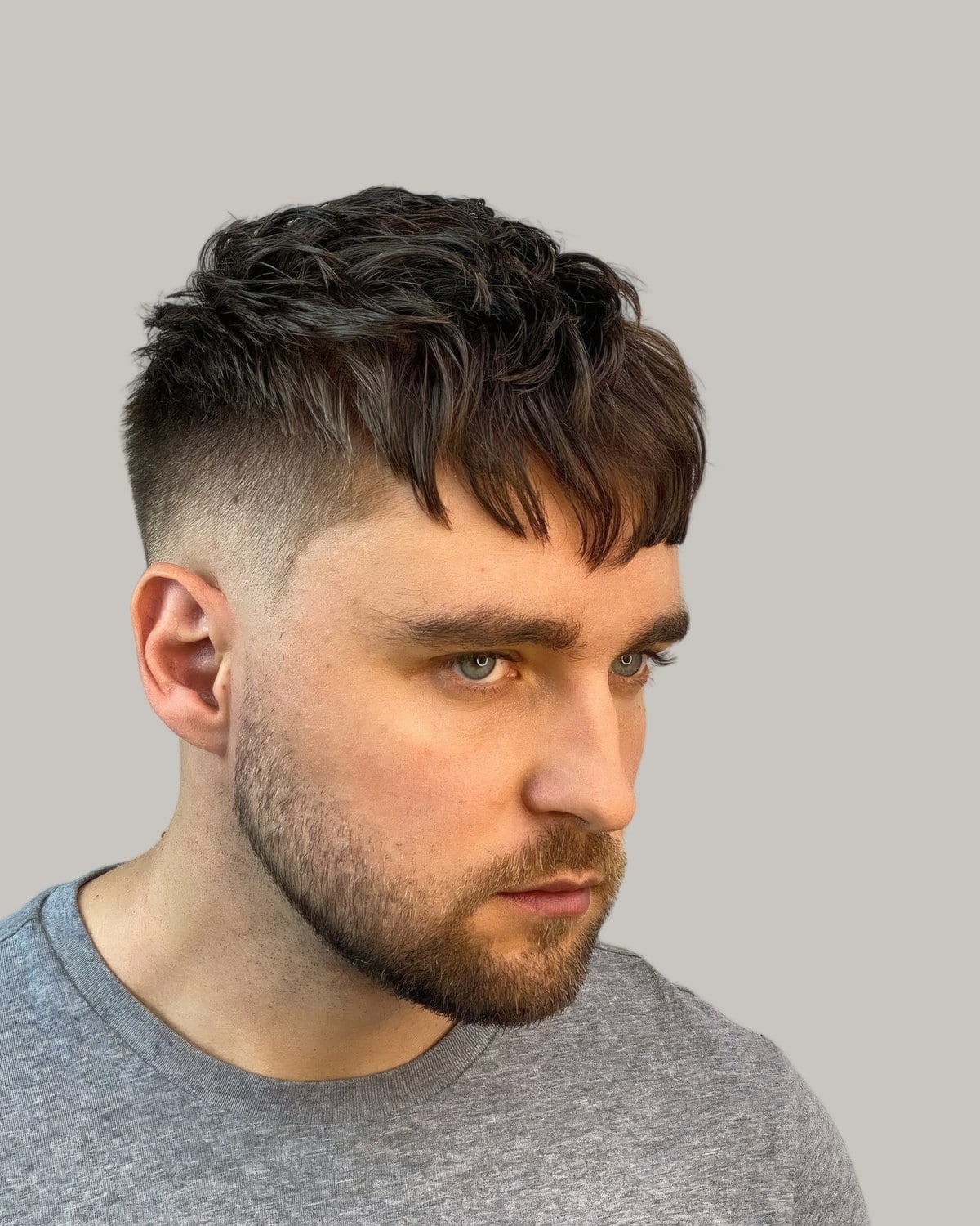 Bearded man in a grey T-shirt with a short textured fringe and a mid fade, looking to the side