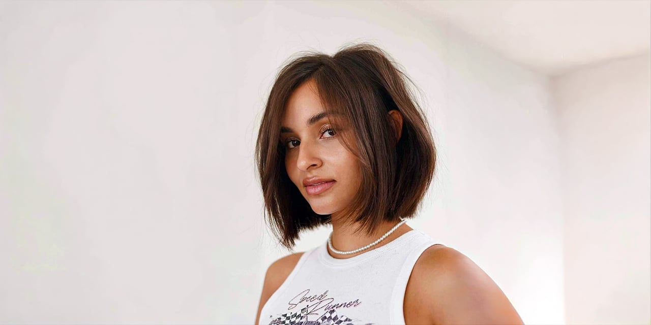 Woman with a brunette blunt bob tucked behind one ear, wearing a pearl necklace and a white tank top