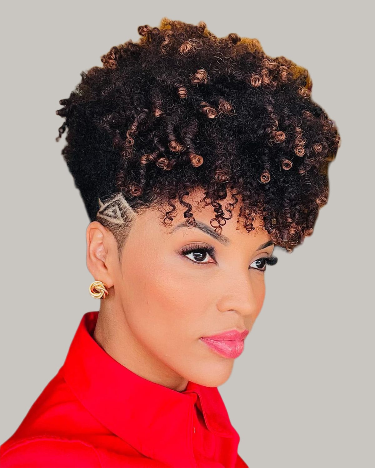 Woman with a tapered cut, a shaved design above the ear and a high crown of black coils with brown highlights, seen in three-quarter view