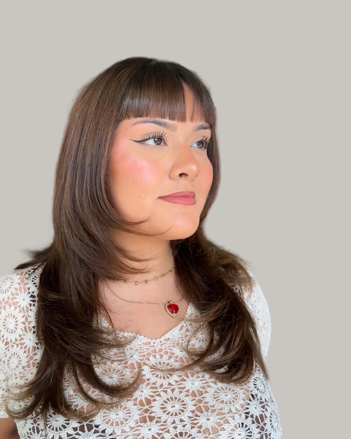 Smiling woman with long brown layers and full bangs, in a white lace top