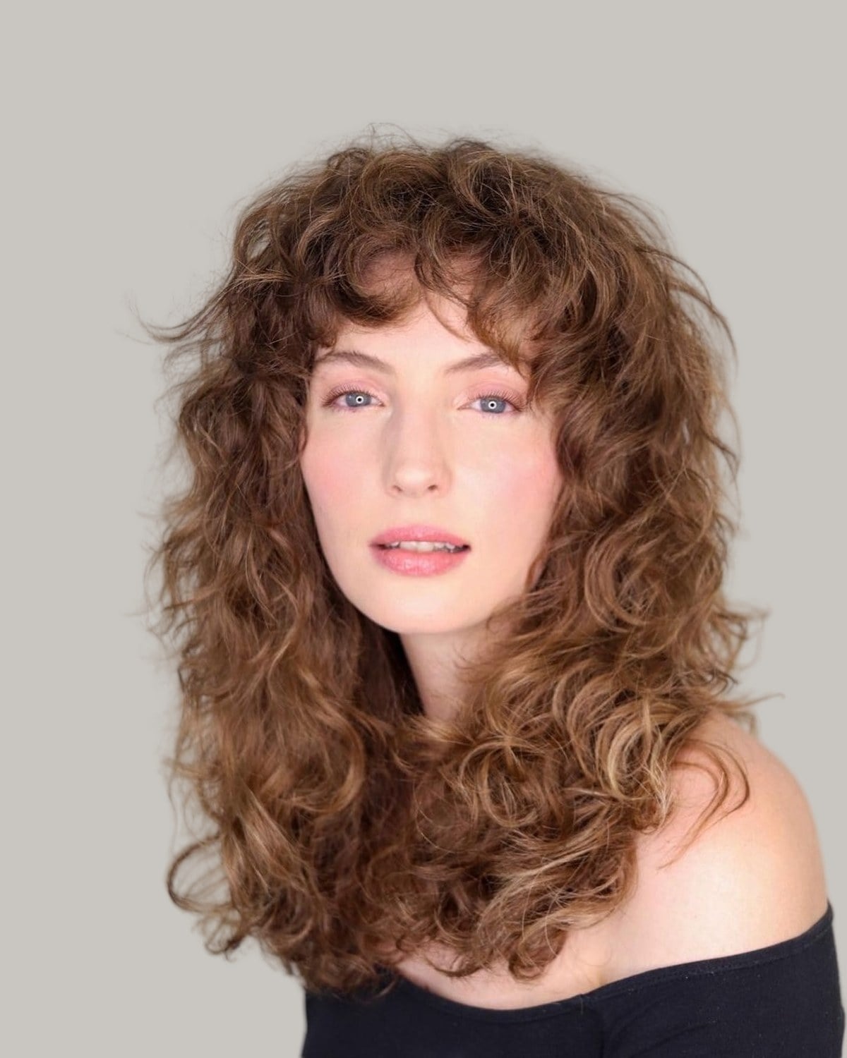 Woman with long auburn curls and curly bangs, in an off-shoulder top