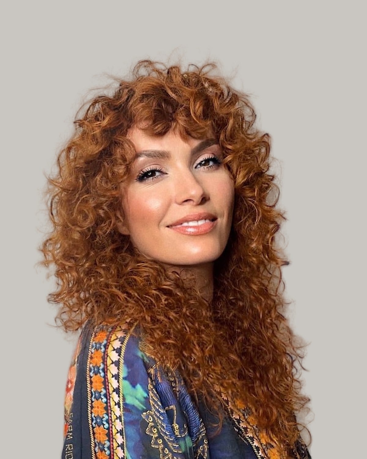 Smiling woman with long copper curls and curly bangs, in a patterned top