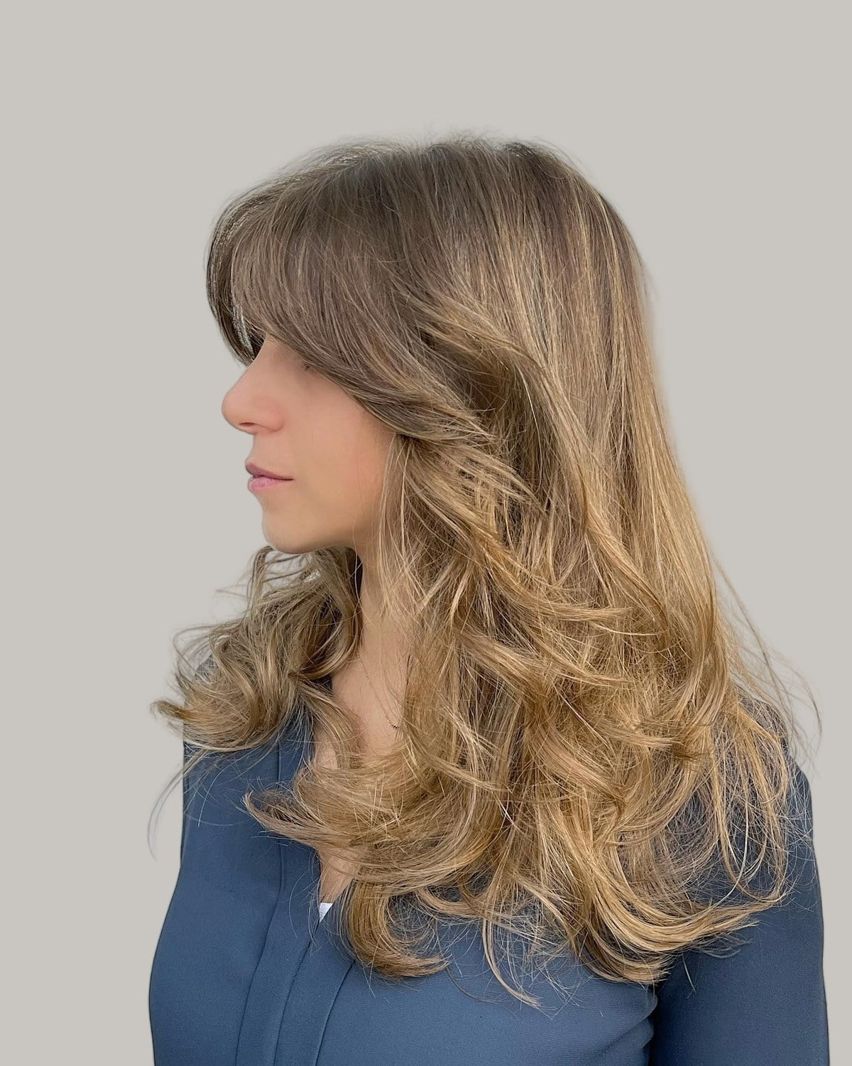 Woman with long, wavy light-brown layers and curtain bangs, seen in profile