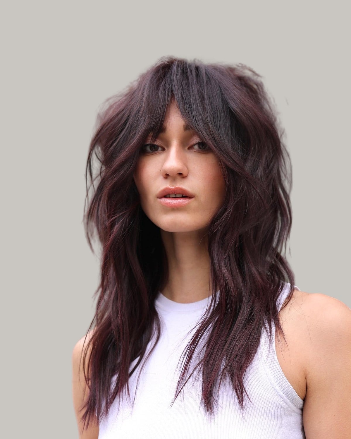 Woman with a long plum-brown wolf cut and bangs, in a white tank top