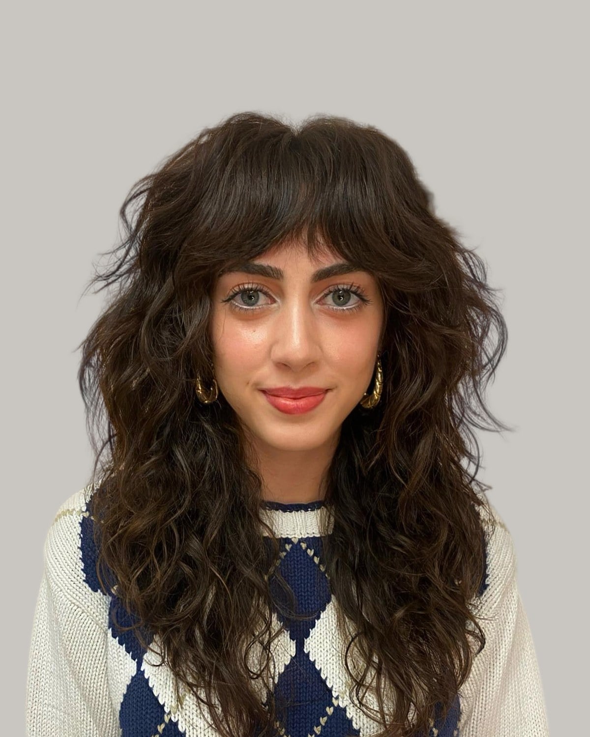 Woman with a long, dark-brown wavy wolf cut and bangs, in an argyle vest